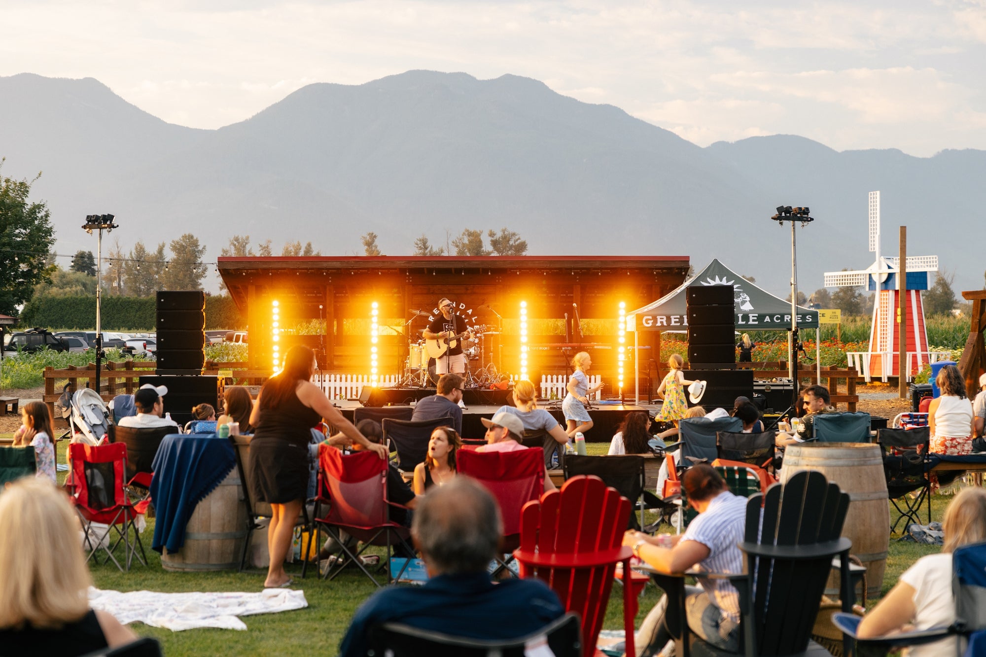 Outdoor concert or event with people seated in chairs and on blankets, stage with lights, and mountains in the background.