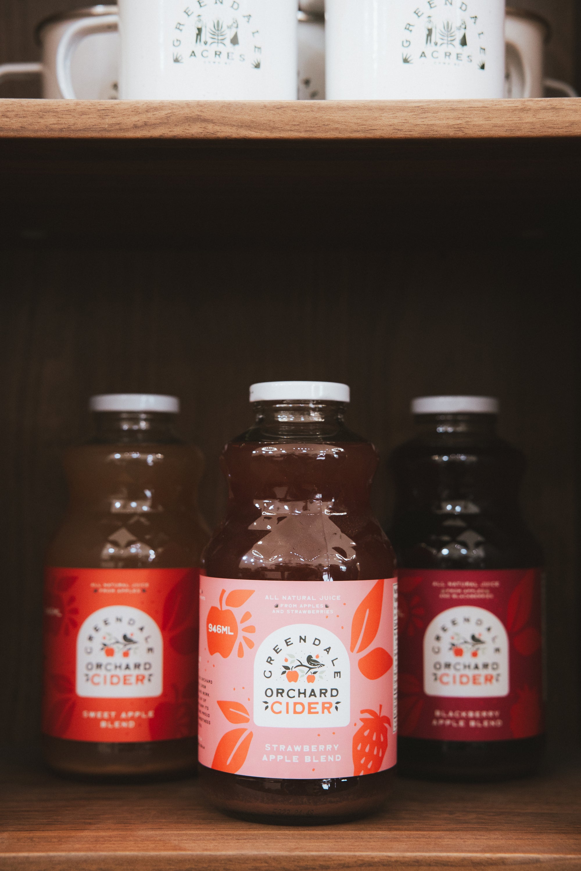 Three bottles of Evernagel Orchard Cider arranged on a wooden shelf, with the center bottle showing a pink label and the side bottles showing red labels.