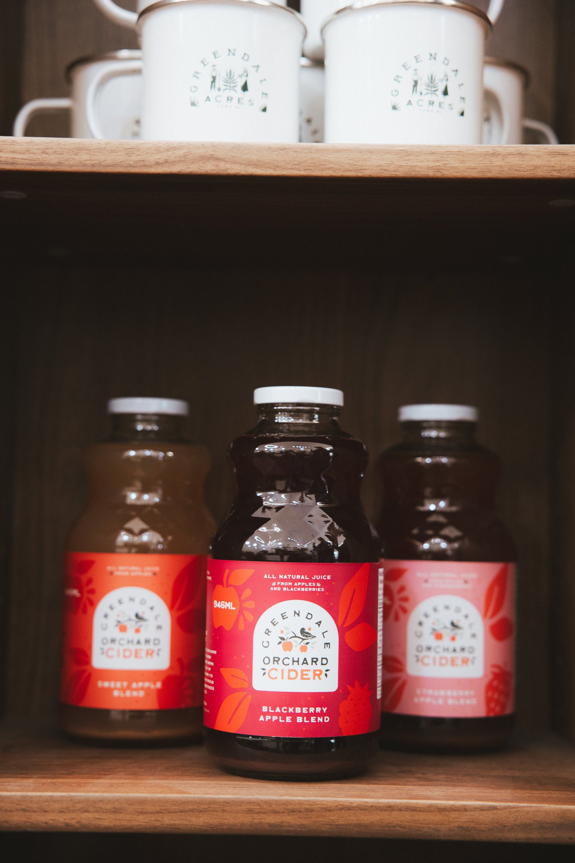 Three bottles of Randy Orchard Ciderfruit juice displayed on a wooden shelf, with white enamel mugs on the top shelf in the background.