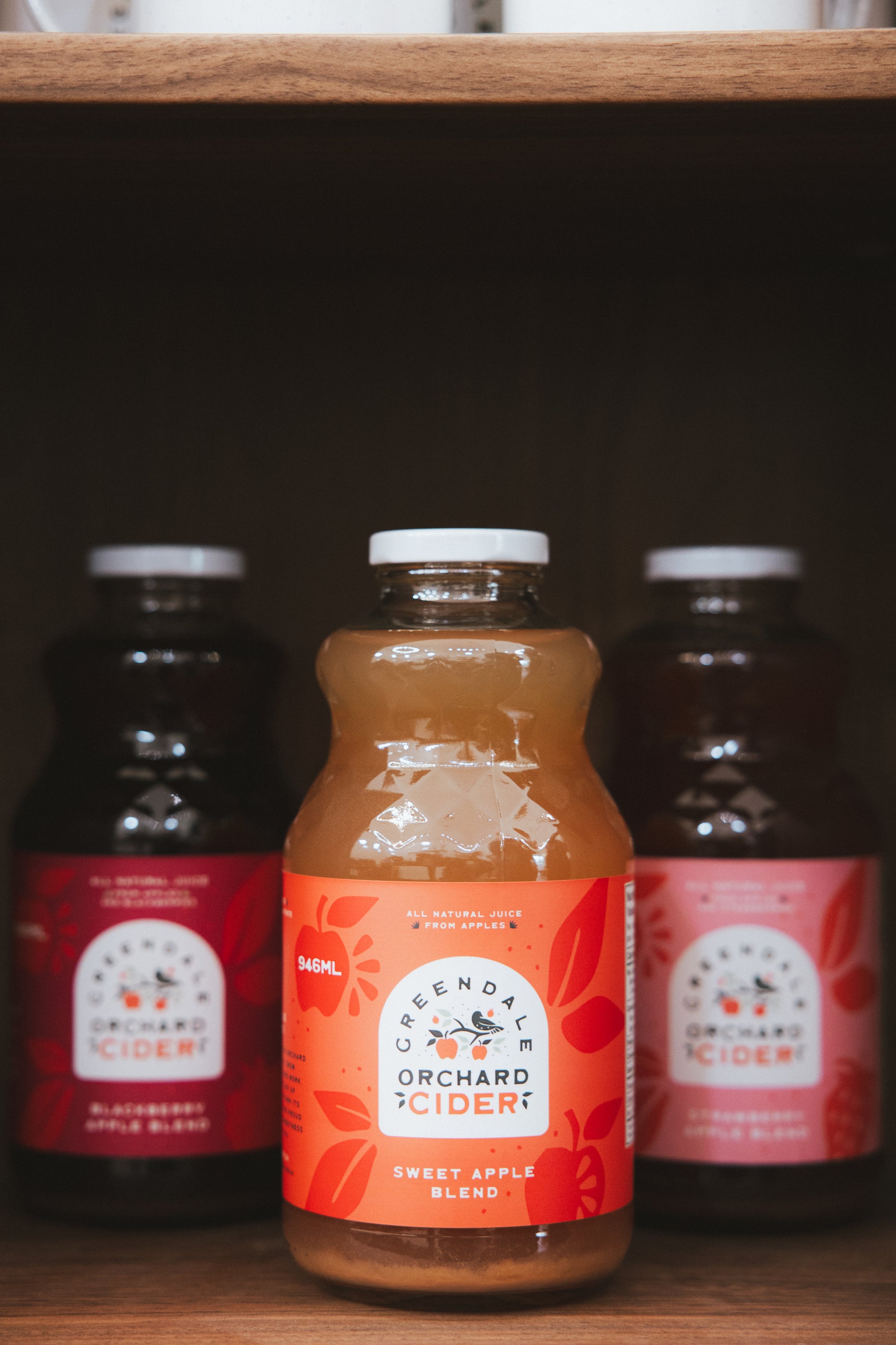 A bottle of Seendag Orchard Sweet Apple Blend cider on a wooden shelf, with two darker bottles in the background.