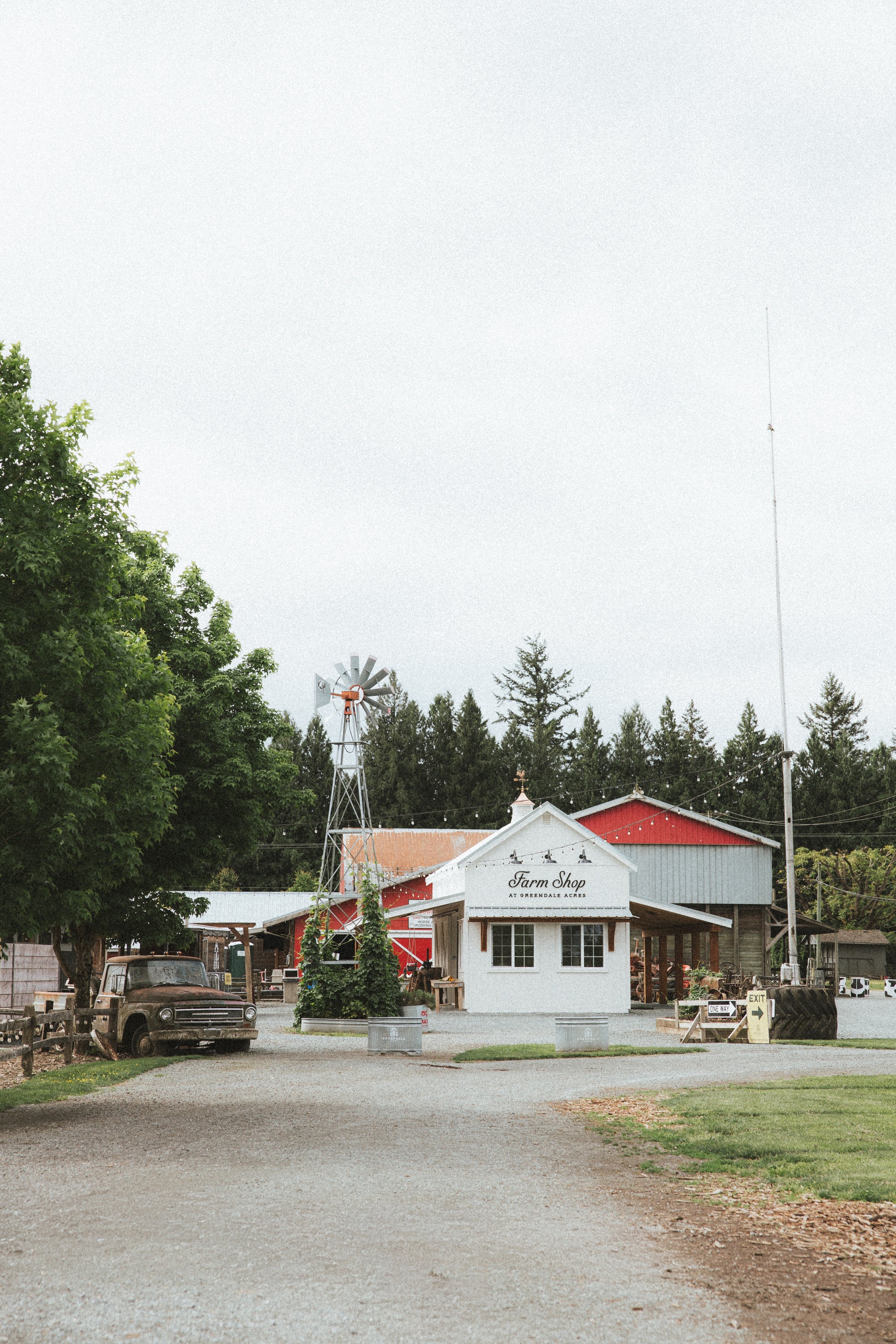 A scenic outdoor photograph of a small white building with red roof sections, a vintage truck parked nearby, a windmill, and tall trees under an overcast sky.