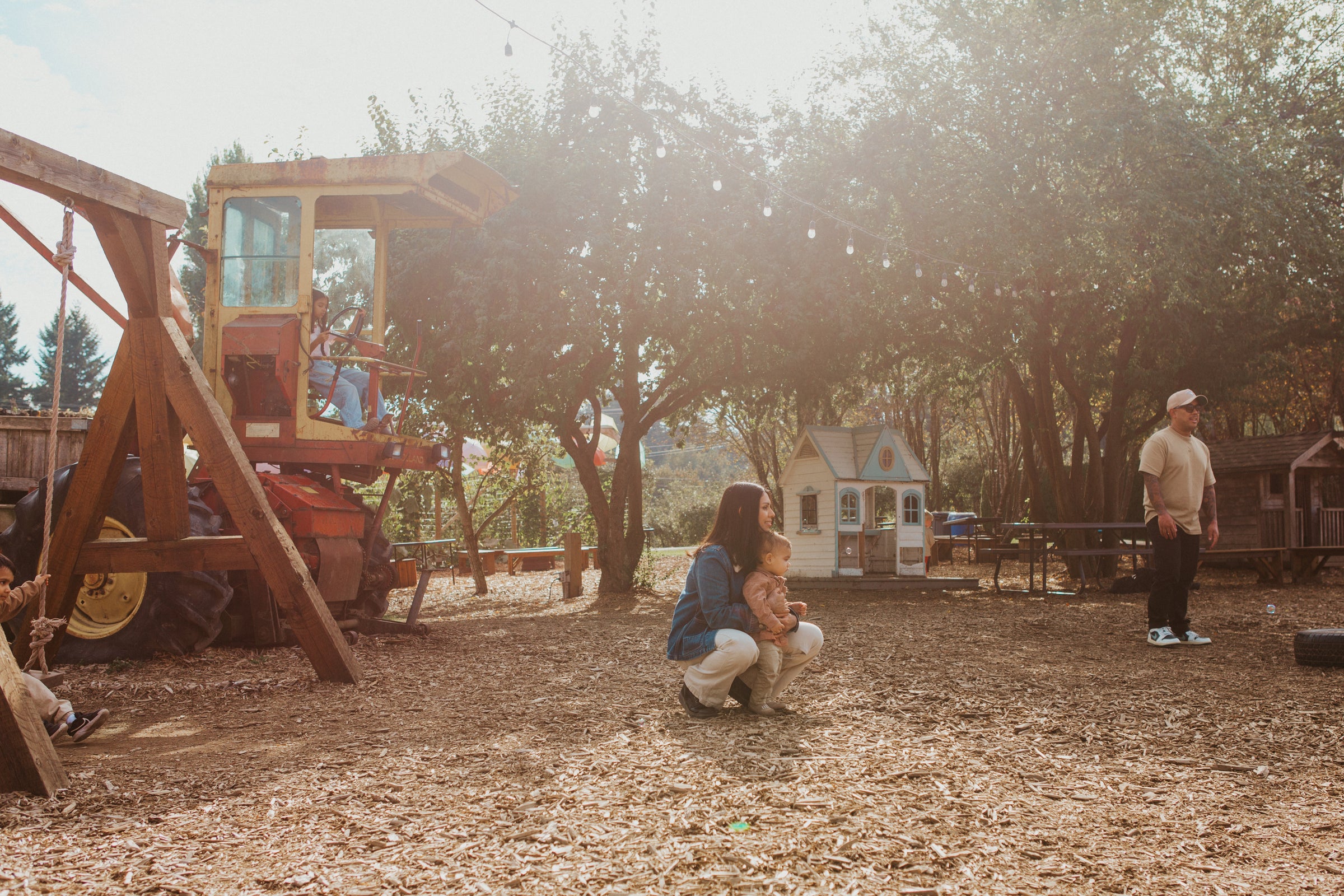 A family scene in a rustic outdoor backyard with a wooden swing set, trees, and a small white playhouse. A woman crouches in the center holding a child, while a man stands to the right.